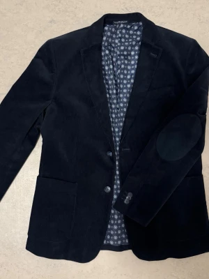 Blå Kavaj från Hampton Republic - Säljer en royalblå/mörkblå kavaj från Hampton Republic i herrstorlek 50. Plagget är helt nytt och aldrig använt, i perfekt skick utan några defekter alls. Kavajen har en stilren, klassisk utsida och ett snyggt mönstrat innerfoder som ger en extra elegant touch. Passar perfekt till både fest, arbete eller mer uppklädda tillfällen.