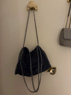 Svart axelväska med kedja - Säljer en svart axelväska med coola silverfärgade kedjor som axelremmar och runt kanten. Väskan har en mjuk form och är tillverkad i mocka. Insidan har ett fack med dragkedja för småsaker. Perfekt storlek för att få plats med allt du behöver.