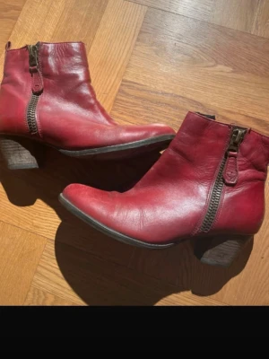 Röda boots i skinn med dragkedja - Snygga röda boots i skinn med rund tå och låg klack. Skorna har en markerad dragkedja på sidan och en robust träfärgad klack. Perfekta för dig som vill sticka ut med färg och gillar klassisk men edgy stil.