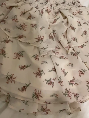 Blommig kortkjol från Gina Tricot - Supersöt kortkjol från Gina Tricot i ljusbeige med små blommor i rosa och grönt. Kjolen har volangdetaljer och smock i midjan för en skön passform. Perfekt för sommarens alla dagar, och materialet är lätt och luftigt.andvändning:0 gånger.stretchig.