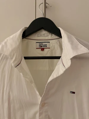 Vit skjorta från Tommy Hilfiger Denim - Vit skjorta från Tommy Hilfiger Denim i slim fit-modell. Klassisk design med knappar framtill, lång ärm och liten broderad logga på bröstet. Skjortan är gjord i bomull och har diskreta rödblå detaljer vid kragen. Perfekt för en clean och stilren look. L passar även M. 