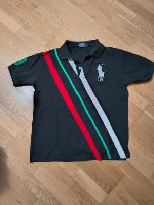 Svart piké från Polo Ralph Lauren - Svart pikétröja från Polo Ralph Lauren med stora diagonala ränder i rött, grönt och grått på framsidan. Klassisk krage med vit text 'RALPH LAUREN' på insidan, korta ärmar och broderad logga på bröstet. Materialet är bomull.
