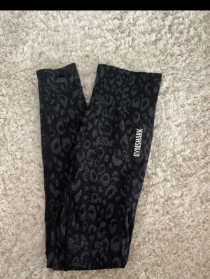 Svarta mönstrade tights från Gymshark - Säljer ett par svarta tights från Gymshark med grått mönster. De har hög midja och är tillverkade i ett stretchigt material som sitter tight och bekvämt. Perfekta för träning och har Gymshark-logga på benet samt text vid höften. Köpta i strl XS, passar även S då de töjer sig lite vid användning.