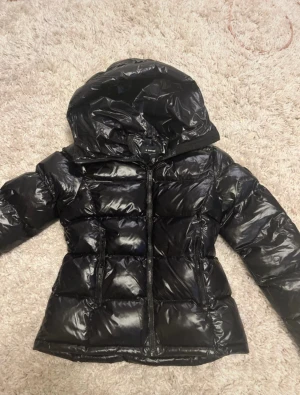 Svart pufferjacka från Everest - Svart glansig pufferjacka från Everest med huva och dragkedja framtill. Jackan har quiltad design och två sidofickor med dragkedja. Perfekt för kalla dagar och har en snygg, modern look som funkar till streetwear.