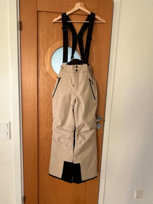 Superfina Beige täckbyxor från Everest, stl 34 - Säljer ett par superfina beige täckbyxor från Everest, stl 34, med svarta justerbara hängslen och svarta detaljer vid benslut och dragkedjor. Byxorna har hög midja, två fickor med dragkedja och förstärkning vid bensluten. Perfekta för vinterns äventyr, endast använda en vecka, jättefint skick, som nya! 