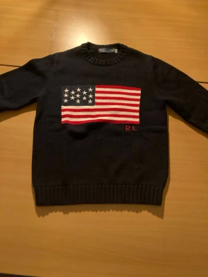Mörkblå stickad tröja Polo Ralph Lauren - Mörkblå stickad tröja från Polo Ralph Lauren med amerikansk flagga i vitt, rött och blått på bröstet. Rund hals och ribbade muddar vid ärmslut och nederkant. RL-broderi i rött under flaggan. Perfekt för dig som gillar klassisk och cool stil.