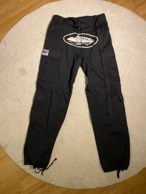 Svarta Cortiez Byxor - Svarta cargopants från Cortiez med flera fickor och ett stort vitt tryck framtill. Byxorna har snörning vid bensluten och en dekorativ rem på ena sidan. Materialet är bomull och byxorna har en loose passform.