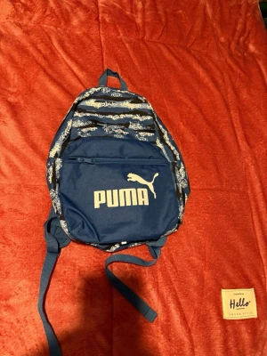 Blå ryggsäck från Puma med mönster - Säljer en blå ryggsäck från Puma med coolt randigt mönster i vitt och svart. Väskan har ett stort fack och en framficka med dragkedja, samt justerbara axelremmar. Perfekt för skola eller häng med kompisar. Puma-logga i vitt på framsidan. Den är liten och passar barn!