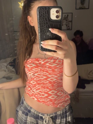 Röd och vit mönstrad tubtopp - Säljer en snygg röd tubtopp med vitt geometriskt mönster. Toppen är axelbandslös och har en bandeau-stil som sitter tight mot kroppen. Perfekt för sommarens varma dagar och enkel att matcha med jeans eller shorts.