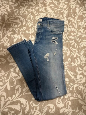 Slim fit blå jeans Glenn 32/32 - Slim fit jeans i klassisk blå tvätt med slitna detaljer och snygga revor på benen. Modellen är smal och sitter tajt längs benen, perfekt för dig som gillar en modern look. Materialet är mjukt jeans och midjan är normal. Passar dig som vill ha en cool och avslappnad stil.