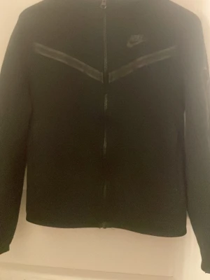 Svart Nike vindjacka med huva - Svart vindjacka från Nike med huva och hel dragkedja framtill. Jackan har diskreta svarta detaljer på bröstet och ärmen, samt en ficka med dragkedja på ena ärmen. Är som gott som ny 