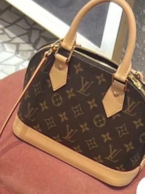 Louis Vuitton 💖 - Säljer en snygg handväska från Louis Vuitton med klassiskt monogrammönster i mörkbrunt och beige. Väskan har ljusa handtag och detaljer i skinn, samt guldfärgade metalldetaljer och dragkedja. Perfekt storlek för det viktigaste och har en stilren, rundad form.