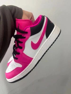Nike Jordan 1 Low rosa och svart - Säljer ett par Nike Jordan 1 Low sneakers i en färgkombo med rosa, svart och vitt. Skorna har klassisk snörning, rund tå och platt sula. Materialet är skinn och syntet, med svart swoosh och detaljer. Perfekt för dig som vill sticka ut med dina outfits.