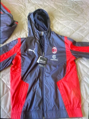 AC Milan vindjacka från Puma - Snygg AC Milan vindjacka från Puma i svart och rött med huva och dragkedja. 