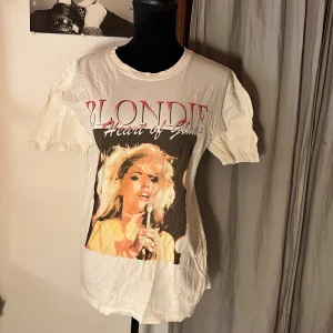 Vit Blondie t-shirt från Pull&Bear - Säljer en vit t-shirt från Pull&Bear med Blondie-tryck och texten 'Heart of Glass' på bröstet. Klassisk passform med korta ärmar och rund hals. Perfekt för dig som gillar bandtröjor och vill ha en cool vintagekänsla.
