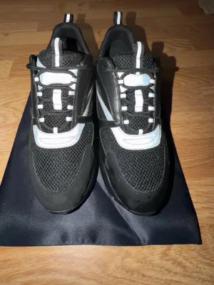 Svarta chunky sneakers med vita detaljer - Säljer ett par svarta chunky sneakers från Dior med vita och grå detaljer. Skorna har meshpartier, snörning och en kraftig plattformssula. Snyggt sportig look med rund tå och kontrasterande paneler i syntet och skinn. Perfekt för dig som gillar statement-skor.