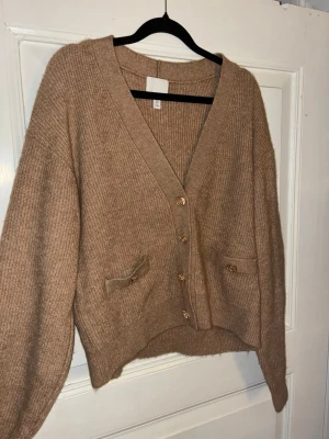 Beige stickad kofta H&M - Säljer en beige stickad kofta från H&M med snygga guldfärgade knappar och två fickor framtill. Koftan har v-ringad hals, långärmade och loose passform som ger en chill vibe. Perfekt att slänga på sig under hösten eller vintern.