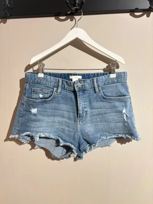 Blå jeansshorts från H&M - Säljer ett par blå jeansshorts från H&M i storlek 38. De har råa, fransiga kanter och slitna detaljer för en avslappnad vibe. Klassisk femficksmodell med knapp och dragkedja framtill. Perfekta till sommaren!