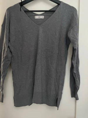 Grå v-ringad tröja från Mango - En enkel och stilren grå tröja från Mango med v-ringning och långa ärmar. Tröjan har ribbade muddar och små slitsar i sidorna för en avslappnad look. Perfekt att matcha med jeans eller kjol.