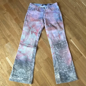 Pastellmönstrade lågmidjade bootcut jeans - Säljer ett par unika jeans från Jaded London med pastellfärgat mönster i rosa, lila och grått. Modellen är bootcut med låg midja och coola detaljer som swirl- och ornamentmönster längs benen. Perfekt för dig som vill sticka ut med din stil.