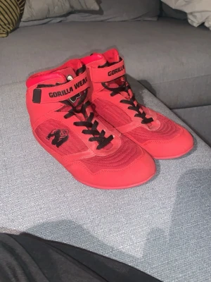 Röda Gorilla Wear träningsskor storlek 42 - Säljer ett par röda träningsskor från Gorilla Wear med svart gorilla-logga på sidan och svarta snören. Skorna har högt skaft, kardborreband upptill och vadderad häl. Materialet är en mix av mocka och syntet, perfekt för gym och styrketräning.
