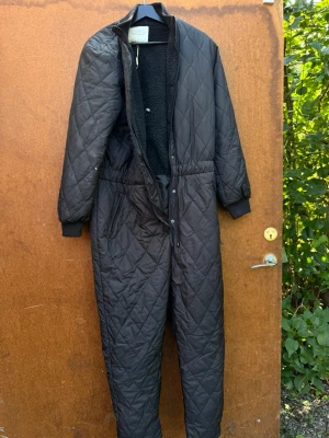 Vinter overall Svart quiltad byxdress från Freequent - Quiltad svart byxdress från Freequent med mysigt teddyfoder på insidan. Byxdressen har dragkedja och knappar framtill, resår i midjan och muddar vid ärmsluten. Perfekt för kalla dagar när du vill vara både varm och stilren.