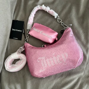 Glittrig rosa axelväska Juicy Couture - • Superfin ljusrosa handväska i strass / kristaller från Juicy Couture. • Väskan kommer med tillhörande portmonnä och ett extra väskrem (axelrem). • Skickas i en vanlig ”dustbag” i satin-liknande tyg. • Helt ny med etikett.