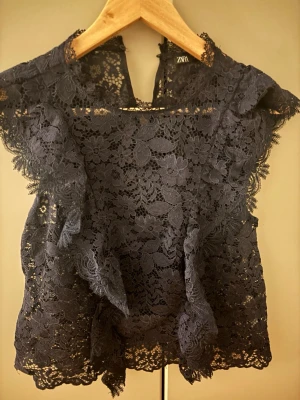 Mörkblå spetsblus från Zara - Supersnygg och trendig mörkblå blus från Zara i genombruten spets med blommigt mönster. Blusen har volangdetaljer längs axlarna och är ärmlös med rund halsringning. Säljs inte längre!! 