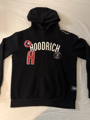 Hoodrich Hoodie - Svart hoodie från Hoodrich med broderade patchar och tryck på bröstet. Stor vit text, röd H och flera detaljerade märken framtill. Klassisk känguruficka och huva med dragsko. Mjuk insida och ribbade muddar. Perfekt för en streetwear-look.