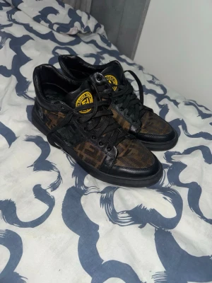 Fendi sneakers med FF-mönster - Säljer ett par snygga Fendi sneakers med klassiskt FF-mönster i brunt och svart. Skorna har svarta skinnpaneler, rund tå och snörning. Fendi-logga i gult på plösen ger en extra lyxig touch. Perfekta för dig som gillar designer och streetstyle.