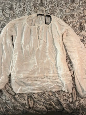 Vit luftig blus med knytning – passar XS–XL - Lätt och luftig vit blus i tunn, mjuk kvalitet med vacker struktur. Blusen har lång ärm, rund halsringning med knytband framtill samt dekorativ spetsdetalj nedtill som ger en bohemisk och feminin känsla. Tack vare den rymliga och avslappnade passformen passar den flera storlekar – från XS till XL. Perfekt både till vardags och sommarens tillfällen. I fint skick.