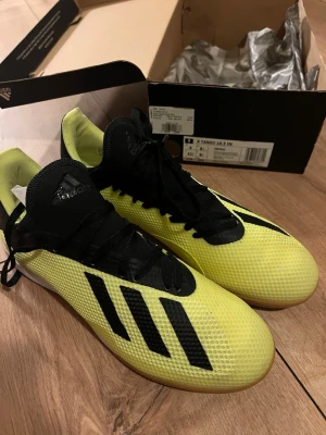Adidas X Tango 18.3 IN fotbollsskor - Säljer ett par Adidas X Tango 18.3 IN inomhusfotbollsskor i svart och gul färg. Skorna har en snygg låg profil, svart snörning och klassiska Adidas-ränder på sidan. Ovandelen är i syntetmaterial för bra bollkontroll och passform. Köpte för flera år sedan, undan lagda och aldrig använda för passade inte mig. 