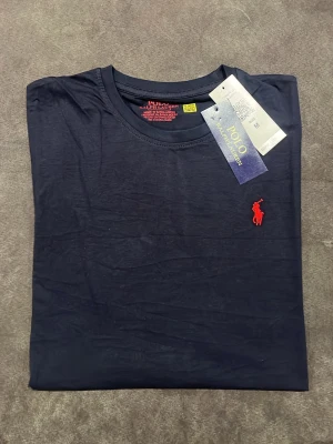 Mörkblå t-shirt från Polo Ralph Lauren - Snygg mörkblå t-shirt från Polo Ralph Lauren med klassisk röd broderad logga på bröstet. T-shirten har rund halsringning och korta ärmar. Tillverkad i mjuk bomull för skön känsla och stilren look.