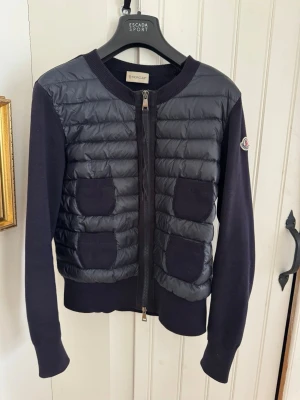 Mörkblå quiltad cardigan jacka Moncler - Snygg mörkblå cardigan jacka från Moncler med quiltad framsida och fyra fickor. Jackan har långärmade stickade ärmar och dragkedja framtill. Materialmix med ylle, nylon och dunfyllning fram. Inköpt på Harrods i London (se kvitto). Sparsamt använd, några maskor i kragen uttänjda (se bilder) annars fint skick. Fler bilder finns på Qr-kod, etiketter, mm 😊