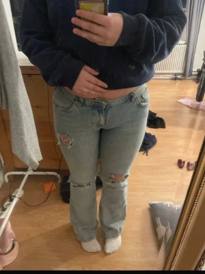 Ljusblå bootcut jeans med slitningar - Säljer ett par ljusblå jeans med bootcut passform och snygga slitningar vid knäna. Jeansen har klassisk femficksdesign och är tillverkade i jeansmaterial. Perfekta för dig som gillar en avslappnad och trendig stil.