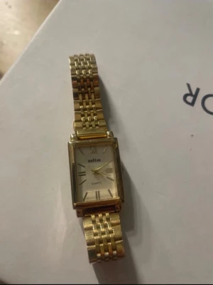 Guldig klocka - Säljer en guldfärgad klocka från Sottas med rektangulär urtavla och metallarmband.