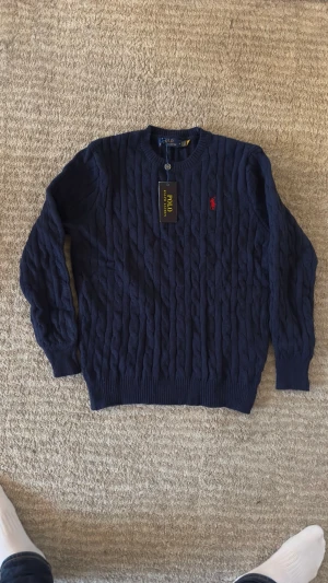 Mörkblå stickad tröja Polo Ralph Lauren - Säljer en mörkblå stickad tröja från Polo Ralph Lauren i storlek M. Tröjan har ett klassiskt kabelstickat mönster och rund hals. På bröstet finns den ikoniska röda Polo-loggan broderad. Perfekt för dig som gillar stilrena och tidlösa plagg.