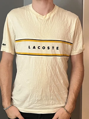 Vit Lacoste t-shirt med gul rand - Säljer en vit t-shirt från Lacoste med korta ärmar och rund hals. Framsidan har en bred gul och svart rand med Lacoste-logga tryckt i mitten. Materialet är mjuk bomull och t-shirten har en klassisk passform. Perfekt för dig som gillar stilrena och sportiga plagg.
