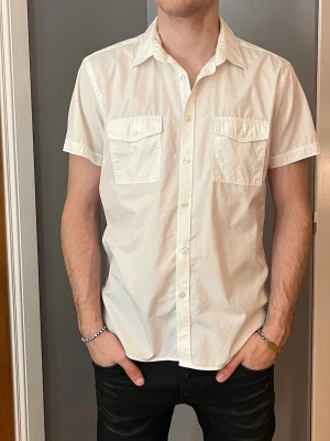 Vit kortärmad skjorta med fickor - Klassisk vit skjorta i bomull med korta ärmar och två bröstfickor med lock och knapp. Skjortan har normal passform och knäppning framtill. Perfekt för en clean och stilren look.