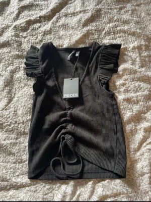 Svart ribbad topp från Pieces - 🖤 Svart ribbad topp med volang – PIECES Storlek: S Skick: Helt ny med prislapp✨  Supersnygg svart ribbad topp från Pieces med volangärmar och knytning framtill. Väldigt smickrande passform och mjukt, stretchigt material. Perfekt både till vardag och fest 🫶  Aldrig använd – prislappen sitter kvar. Lätt att styla med jeans, kjol eller kostymbyxor.  ✔️ Trendig ✔️ Ny med tags ✔️ Stretchig & bekväm  Skicka gärna meddelande vid frågor eller intresse 💌