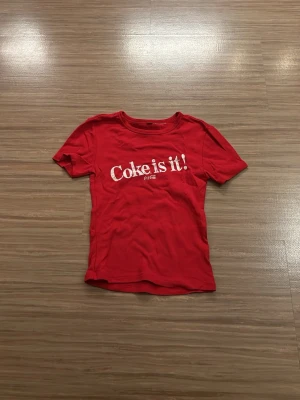 Röd Coca-Cola t-shirt med tryck - Röd t-shirt från Coca-Cola med det klassiska trycket 'Coke is it!' framtill. T-shirten har rund halsringning och korta ärmar. Perfekt för dig som gillar retrostil och vill sticka ut med en ikonisk logga.Utan lapp men är från H&M