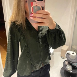 Mörkgrön blus med volangdetaljer - Säljer en mörkgrön blus med liten v-ringning och knappar framtill. Blusen har volangdetaljer längs sidorna💝Bilderna är från förra säljaren, skicka för fler bilder💕