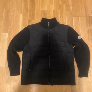 Svart Moncler pufferjacka med stickade ärmar - Säljer en svart pufferjacka från Moncler med ribbstickade ärmar och hög krage. Jackan har dragkedja framtill och två fickor. Klassisk Moncler-logga på ena ärmen. Perfekt för dig som vill ha en stilren och varm jacka med snygga detaljer. Xs