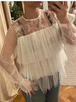 Snyggaste volangblusen💕 - Superfin vit blus i transparent mesh med långa ärmar, volanger och små pärlor. Överdelen har broderade blommor i spets och knytning bak i nacken. Perfekt för dig som gillar romantisk och drömmig stil. Säljes till bästa pris! Liknar Ida Sjöstedt blusen. Se sista bild för den lilla defekten men syns knappt när man har på sig den då volangerna täcker det💕