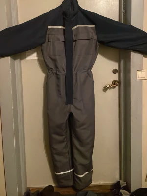 Grå täckbyxa/overall från Powerfix - Säljer en grå och mörkblå täckbyxa/overall från Powerfix med reflexdetaljer vid benen och bröstfickor med lock. Tillverkad i slitstarkt syntetmaterial, perfekt för kalla dagar eller arbete utomhus. Dragkedja framtill och resår i midjan för skön passform.