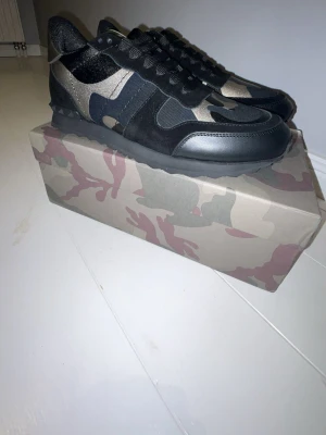 Valentino Rockrunner sneakers camouflage - Säljer ett par Valentino Rockrunner sneakers med svart och grått camouflage-mönster. Skorna har snörning, platt sula och rund tå. Materialmix av skinn, mocka och textil. Snygga detaljer med mörka toner och coola nitar på hälen. Perfekt för dig som gillar streetstyle.