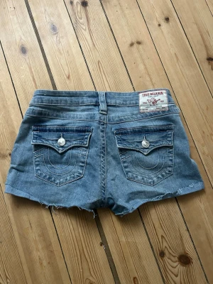 True Religion shorts - Säljer ett par blåa denimshorts från True Religion, modell Joey Low Rise. Shortsen har låg midja och snygga detaljer med knappar och broderade bakfickor. Även avklippt kant nertill.