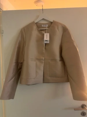 Beige kavaj från Wera, storlek 38 - Snygg beige kavaj från Wera med rund halsringning och två stora fickor framtill. Jackan har en stilren och minimalistisk look med raka, långärmade ärmar och croppad passform. Perfekt för dig som gillar clean och modern stil.