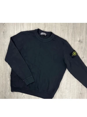Svart sweatshirt från Stone Island - Svart långärmad sweatshirt från Stone Island med klassisk rund hals och ribbade muddar. Ikonisk Stone Island patch med gul brodering sitter på vänster ärm. Perfekt för dig som gillar streetwear och vill ha en clean look.