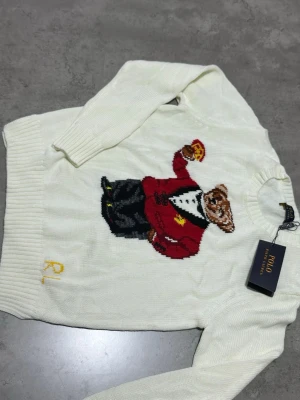 Vit stickad tröja Polo Ralph Lauren - Vit stickad tröja från Polo Ralph Lauren med ikonisk björn i röd kavaj broderad på bröstet. Tröjan har rund halsringning, ribbade muddar och RL-broderi i gult nedtill. Perfekt för dig som vill ha något klassiskt men ändå lekfullt.